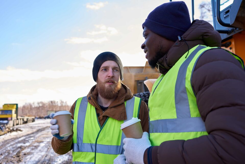 Two workers wearing warm clothing and high-visibility vests discuss ventilation assessment strategies to mitigate cold stress risks at a construction site