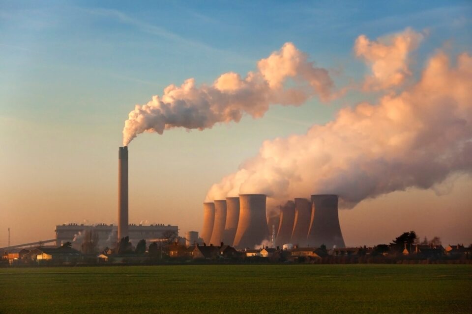 Industrial facility releasing large plumes of smoke from cooling towers and smokestacks during an emissions test, highlighting air pollution levels over a nearby residential area and green field.