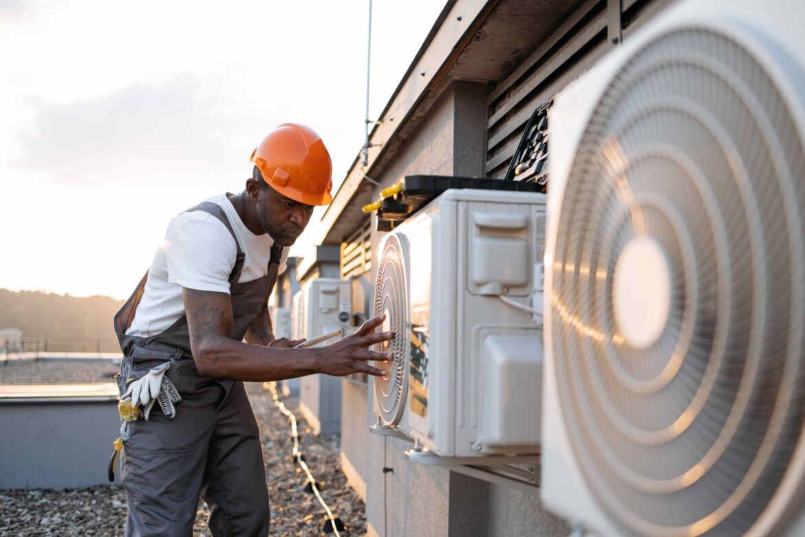 Technician wearing a hard hat and overalls inspecting an outdoor air conditioning unit on a rooftop during sunset, performing a ventilation assessment.