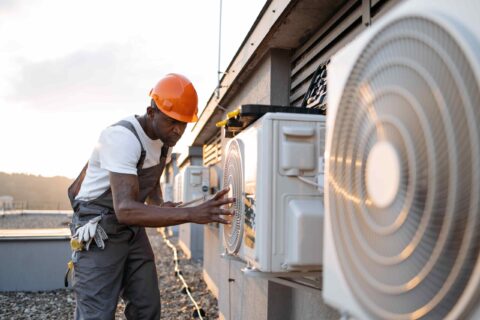 Technician wearing a hard hat and overalls inspecting an outdoor air conditioning unit on a rooftop during sunset, performing a ventilation assessment.